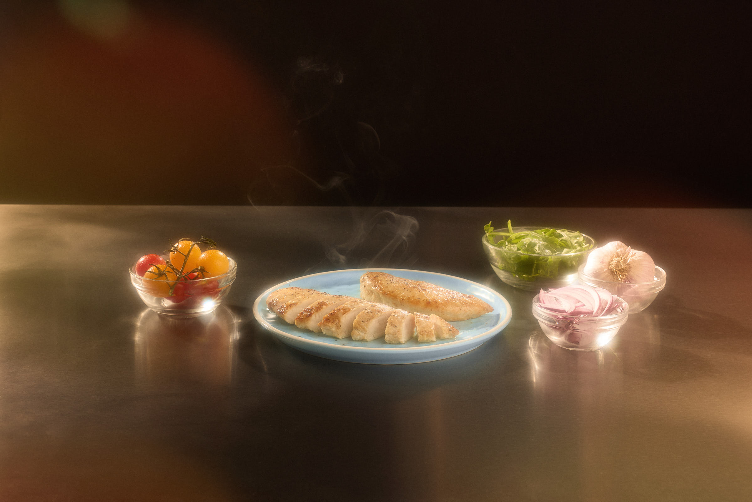Sliced chicken breast on a blue plate, with steam rising beside tomatoes and greens